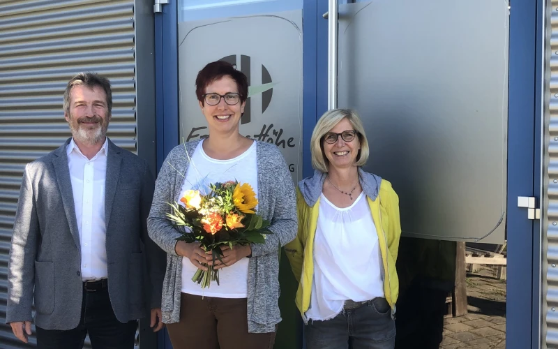 Drei Personen stehen vor einer T&uuml;r, die mit dem Logo der Erlacher H&ouml;he versehen ist. Die Frau in der Mitte h&auml;lt einen Blumenstrau&szlig; in den H&auml;nden. Alle drei l&auml;cheln in die Kamera.