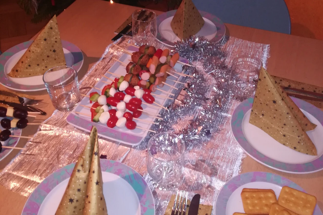 Ein festlich eingedeckter Tisch mit Weihnachtsbaum im Hintergrund ist zu sehen. Eine silberne Lametta-Girlande, ein Teller mit K&auml;secrackern und Tomate-Mozzarella-Spie&szlig;en stehen auf dem Tisch.