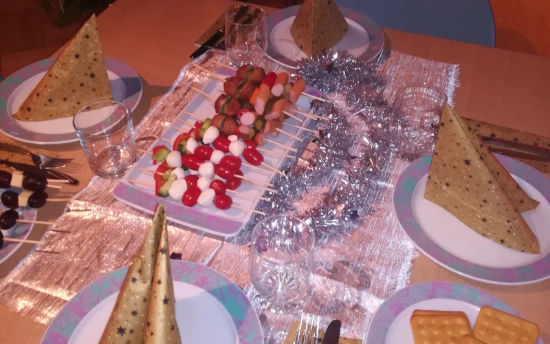 Ein festlich eingedeckter Tisch mit Weihnachtsbaum im Hintergrund ist zu sehen. Eine silberne Lametta-Girlande, ein Teller mit K&auml;secrackern und Tomate-Mozzarella-Spie&szlig;en stehen auf dem Tisch.