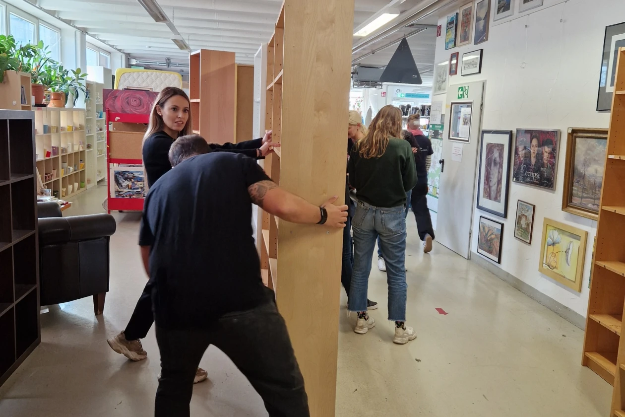 Zwei Personen tragen gemeinsam ein gro&szlig;es Holzregal durch einen Raum mit Regalen und Kunstwerken an den W&auml;nden. Sie bewegen sich vorsichtig mit der sperrigen Ladung.