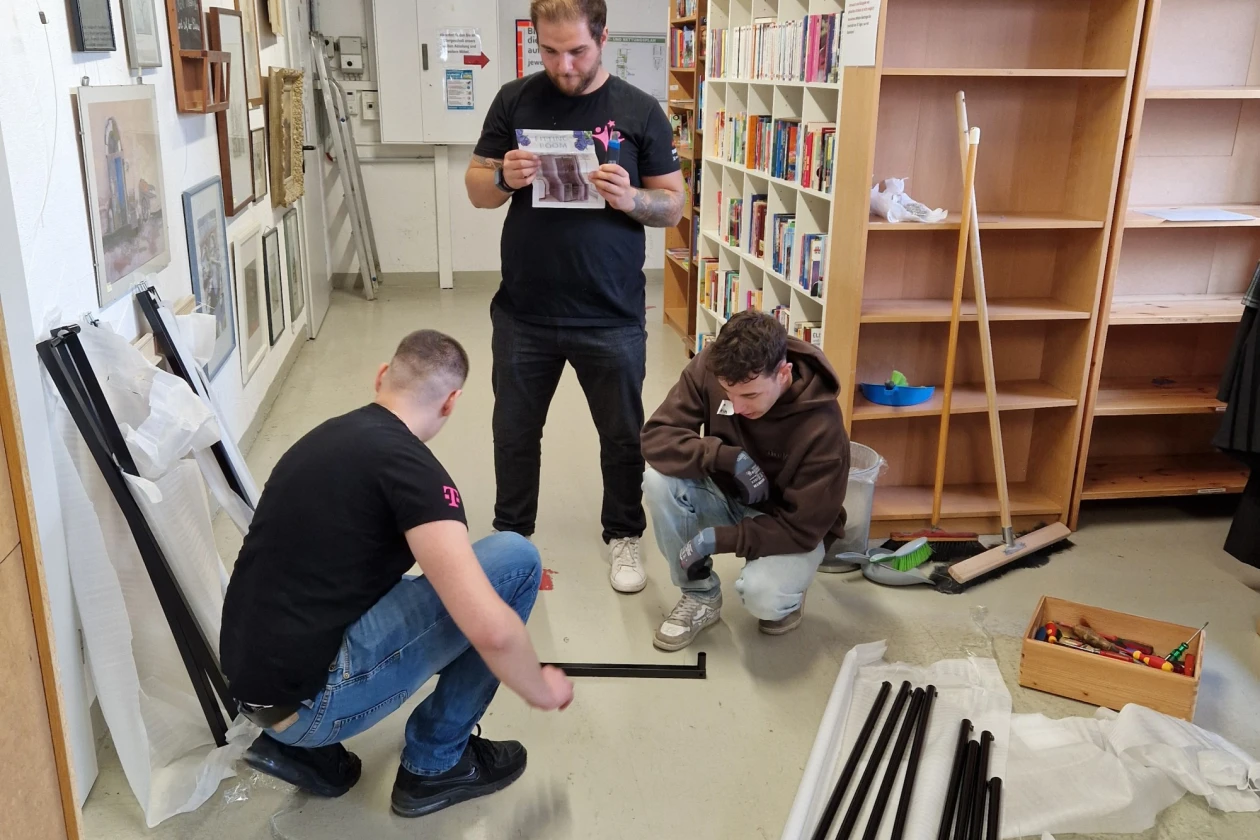 Drei Personen bauen in einem Ausstellungsraum B&uuml;cherregale auf. Sie arbeiten konzentriert und tragen schwarze T-Shirts. Verschiedene Werkzeuge und Metallstangen liegen auf dem Boden verstreut.