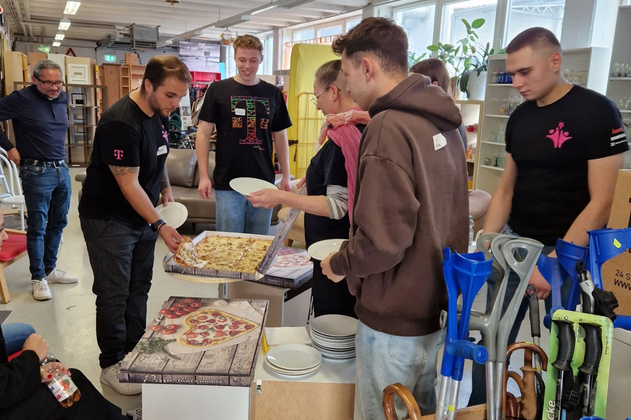 Mehrere Personen stehen an einem Tisch mit Pizza und anderen Speisen in einem hellen Raum Regalen im Hintergrund. Sie scheinen eine Pause mit einer gemeinsamen Mahlzeit zu machen, w&auml;hrend sie in schwarzen T-Shirts gekleidet sind.