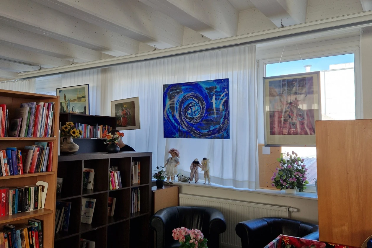 Gem&uuml;tlicher Raum mit einer gro&szlig;en B&uuml;cherwand, einem blauen Kunstgem&auml;lde an der Wand, einem Holztisch mit Blumen und zwei dunklen Sesseln. Wei&szlig;e Deckenbalken und helle Vorh&auml;nge verleihen dem Raum eine freundliche, einladende Atmosph&auml;re.