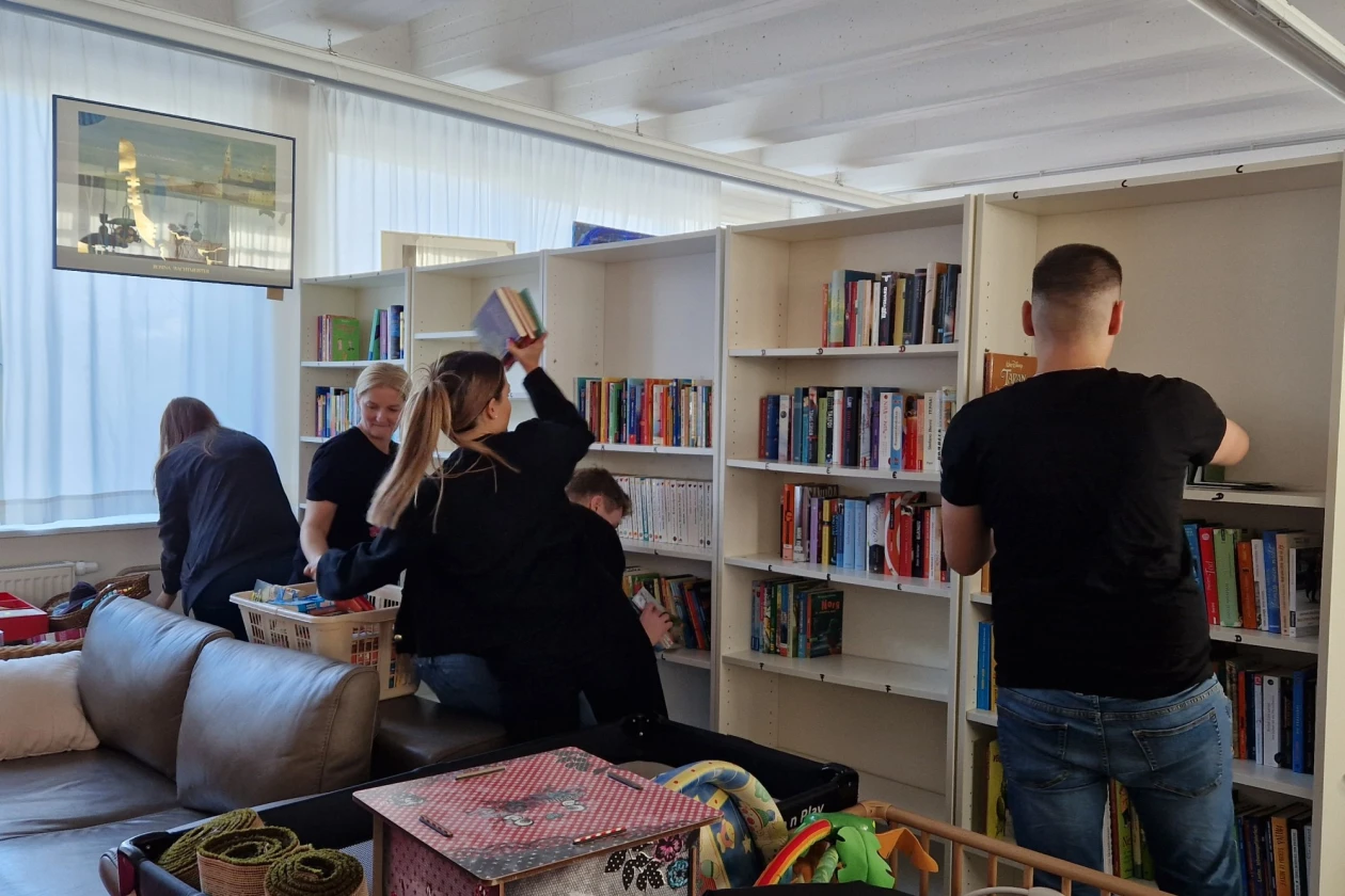 Mehrere Personen in einem hellen Raum mit wei&szlig;en B&uuml;cherregalen, Sofa und gro&szlig;em Fenster, die gemeinsam ordnen und einr&auml;umen. Ein dekoratives Objekt mit einer Illustration ist im Vordergrund sichtbar.