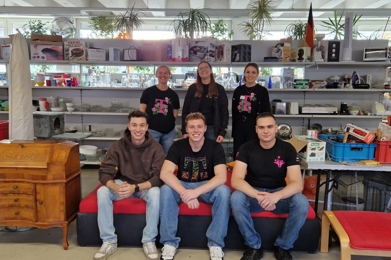 Sechs junge Menschen posieren in einem Sozialkaufhaus und l&auml;cheln in die Kamera. Drei sitzen auf einer roten Bank im Vordergrund und drei stehen im Hintergrund. Alle tragen schwarze T-Shirts mit einem &auml;hnlichen Logo. Der Raum ist modern eingerichtet mit Holzm&ouml;beln, Regalen und gro&szlig;en Fenstern.