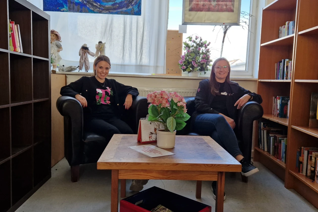 Zwei Personen sitzen in schwarzen Ledersesseln an einem Tisch vor einer Fensterfront. Seitlich stehen B&uuml;cherregale, in den Fenstern h&auml;ngen Kunstwerken. Unter dem Holztisch steht ein roter Rollcontainer.