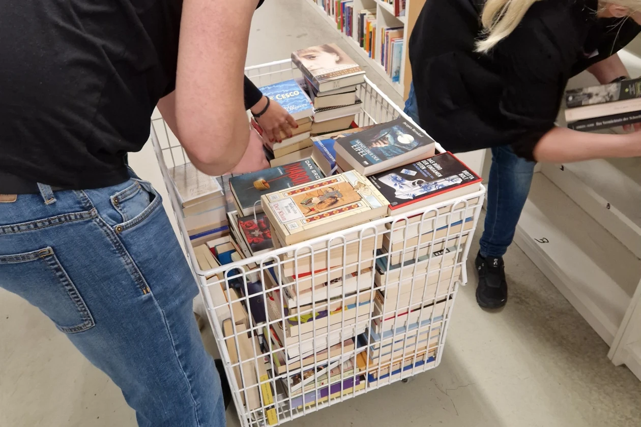 Zwei Personen stehen an einer B&uuml;cherbox und durchsuchen diese in einem Laden. Sie tragen Alltagskleidung und scheinen konzentriert B&uuml;cher zu sortieren und einzur&auml;umen.