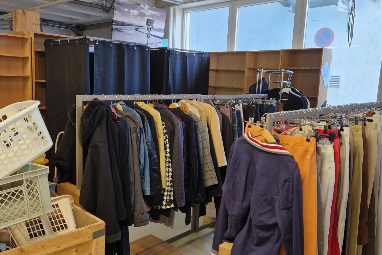 Kleiderst&auml;nder in einem hellen Raum mit verschiedenfarbigen Jacken und M&auml;nteln, umgeben von Holzkisten und Regalen. Gro&szlig;e Fenster lassen Tageslicht herein und beleuchten den Bereich mit verschiedenen Kleidungsst&uuml;cken in gelben, grauen und blauen T&ouml;nen.