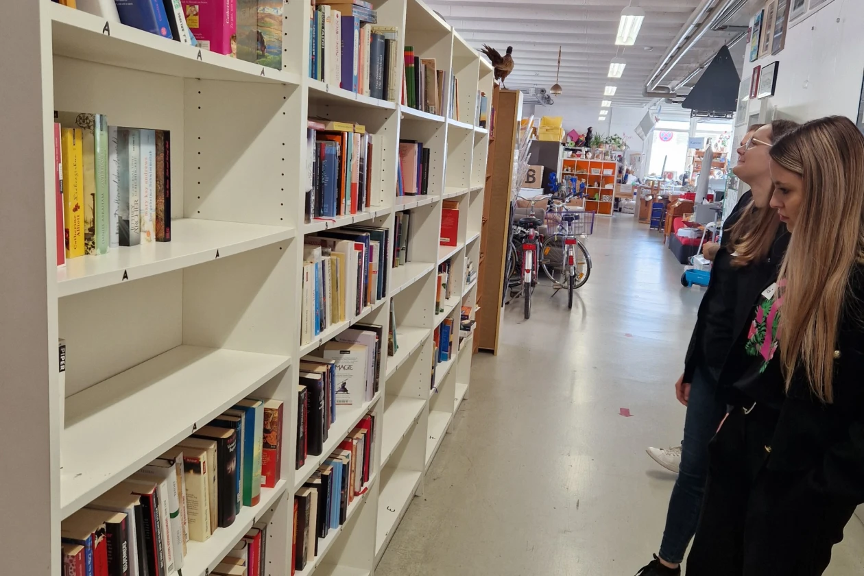 Zwei Personen stehen vor einem B&uuml;cherregal in einem Laden mit Regalen voller B&uuml;cher in verschiedenen Farben. Der Gang ist hell erleuchtet und f&uuml;hrt zu einem Bereich mit Fahrr&auml;dern und anderen Gegenst&auml;nden im Hintergrund.