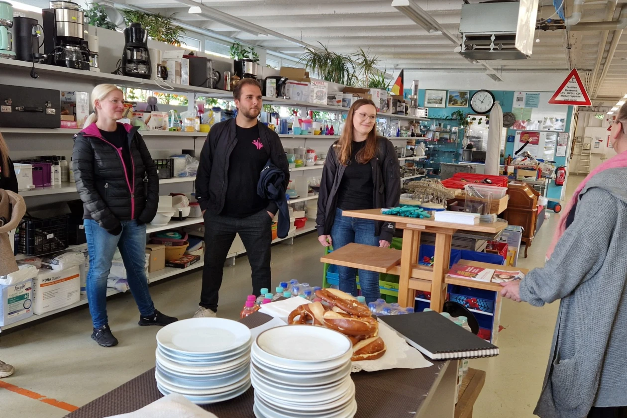 Drei Personen stehen in einem Laden Regalen voller K&uuml;chenger&auml;ten, Geschirr und Glasvasen. Auf einer Theke im Vordergrund stehen Teller und Brezeln. Die Atmosph&auml;re wirkt entspannt und informell.