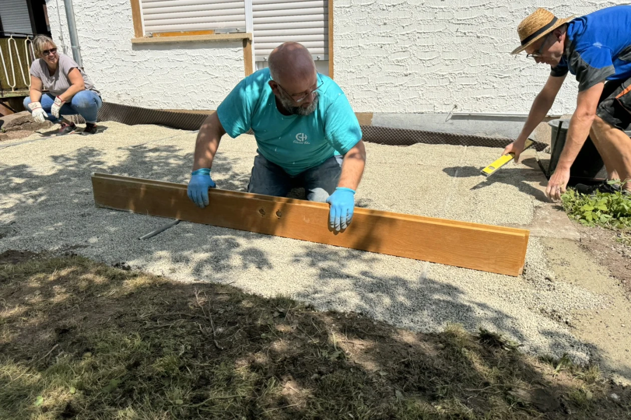 Drei Arbeiter bei der Bodengestaltung: Ein Mann in t&uuml;rkisfarbenem T-Shirt richtet ein Holzbrett auf dem Boden aus um einen ebenen Untergrund f&uuml;r die neue Terrasse abzuziehen. Zwei weitere Kollegen im Hintergrund unterst&uuml;tzen ihn.