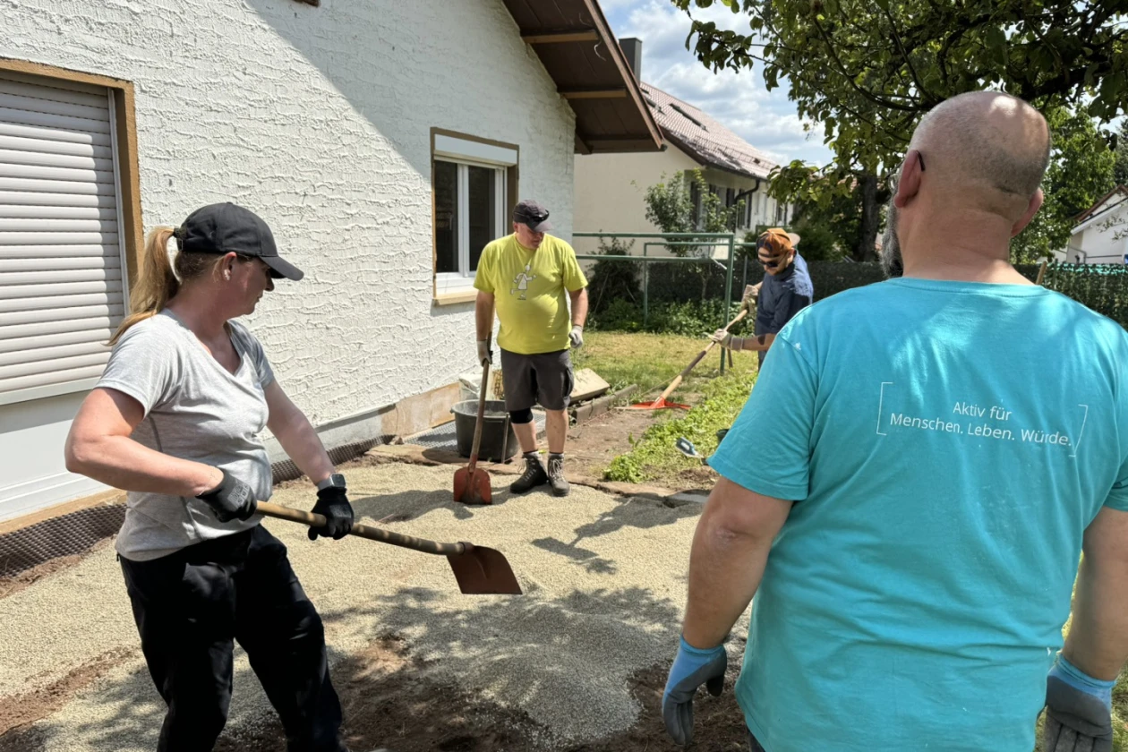Vier Personen beim Arbeiten: Eine Frau mit Schaufel, ein Mann in gelbem T-Shirt, eine Person mit Hut und ein Mann in t&uuml;rkisblauem T-Shirt arbeiten gemeinsam an der Neuerrichtung einer Terrasse an einem Haus.