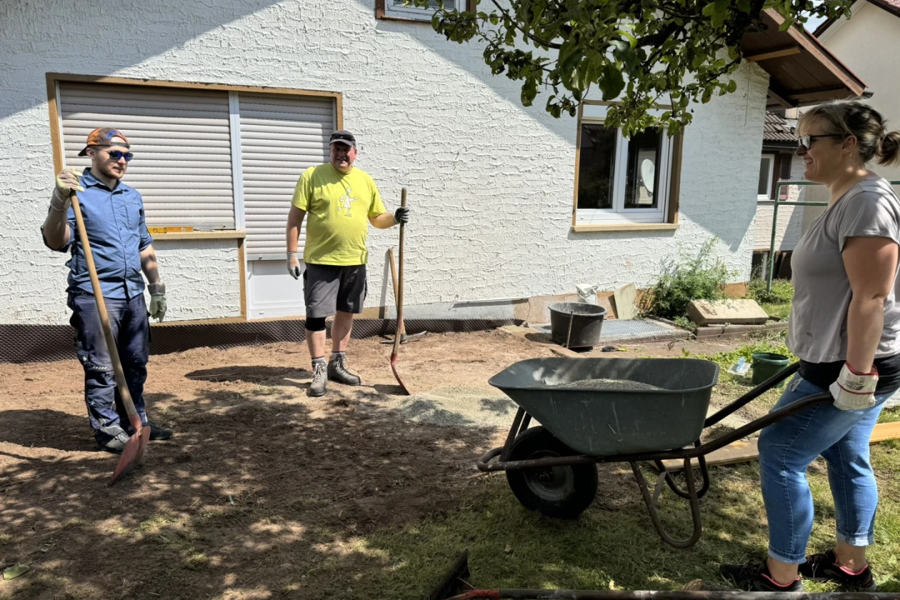 Drei Personen beim Arbeiten mit Schubkarre, Harke und Schaufel vor einem wei&szlig;en Haus an einem sonnigen Tag, besch&auml;ftigt mit der Neugestaltung der Terrasse im Au&szlig;enbereich.