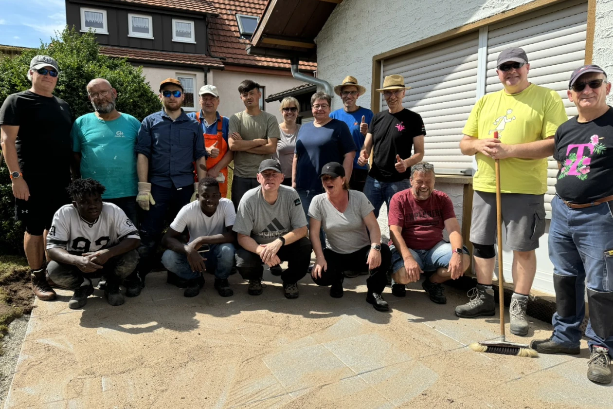 Eine Gruppe Menschen steht f&uuml;r ein Gruppenbild vor der Kamera. Im Hintergrund ist ein Geb&auml;ude zu sehen. Sie stehen auf einer neu mit Platten ausgelegten Terrasse, auf der der Fugensand noch zu sehen ist.