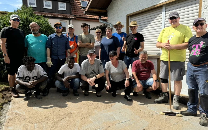 Eine Gruppe Menschen steht f&uuml;r ein Gruppenbild vor der Kamera. Im Hintergrund ist ein Geb&auml;ude zu sehen. Sie stehen auf einer neu mit Platten ausgelegten Terrasse, auf der der Fugensand noch zu sehen ist.