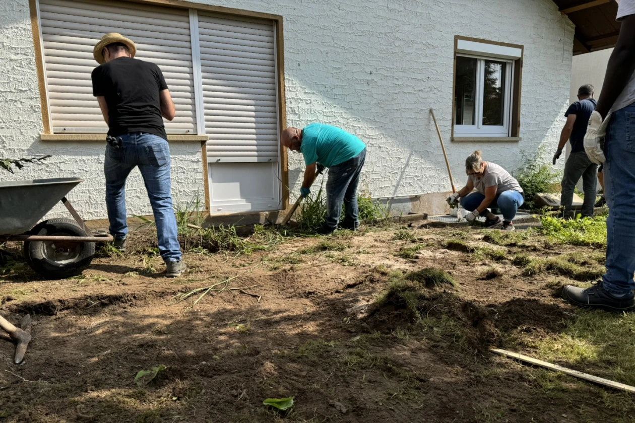 Mehrere Personen arbeiten gemeinsam an der Umgestaltung eines Gartenbereichs, indem sie Erde umgraben und mit Schubkarre und Gartenwerkzeugen die Fl&auml;che vorbereiten.