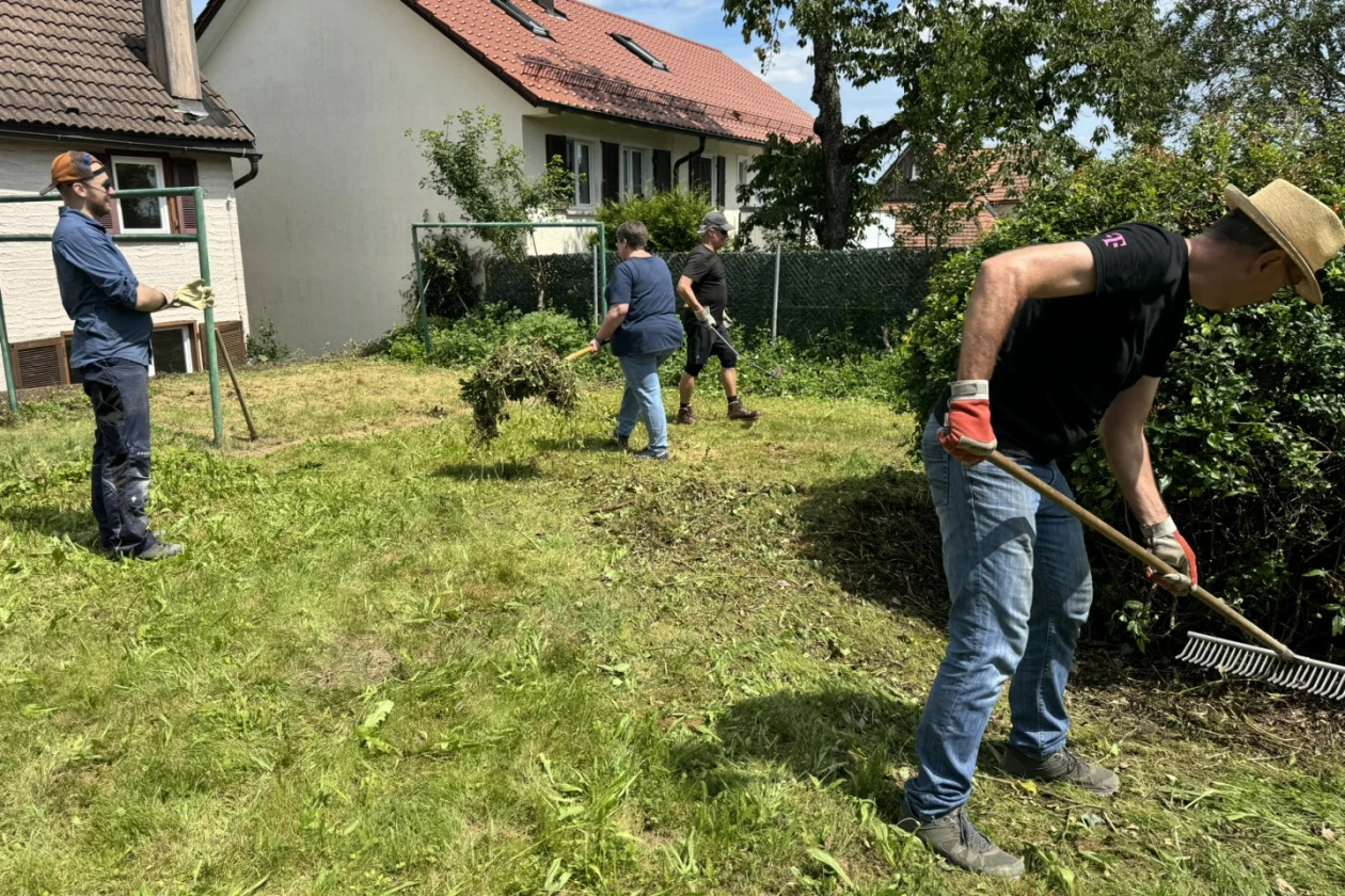 Drei Personen bei der Gartenarbeit auf einem verwilderten Rasengrundst&uuml;ck. Sie m&auml;hen das hohe Gras, rechen Grasschnitt zusammen und s&auml;ubern den Garten. Im Hintergrund sind H&auml;user und B&auml;ume zu sehen.