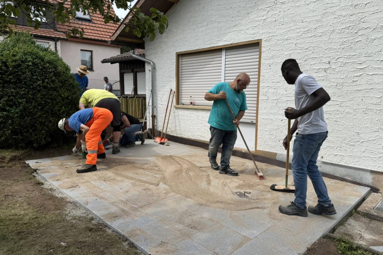 Vier M&auml;nner fugen die Platten einer neuen Terrasse mit Sand aus. Im Hintergrund sind zwei Geb&auml;ude zu sehen uns zwei weitere Personen.