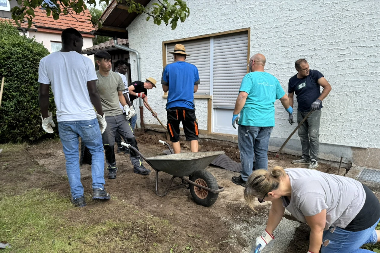 Eine Gruppe von Personen arbeiten gemeinsam an der Umgestaltung in einem Garten. An der Terrassenfl&auml;che wird der Untergrund mit Schaufeln ausgehoben und mit Schubkarre weggefahren.