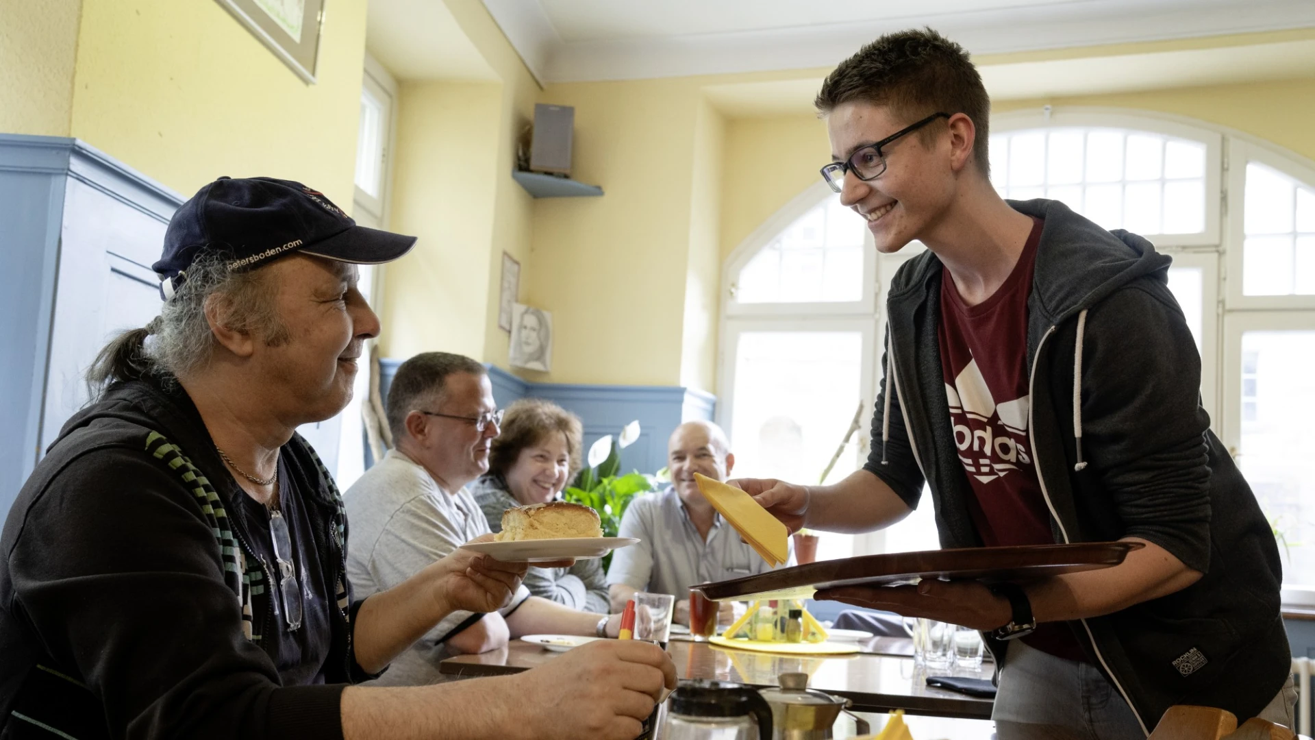 Junger Mann serviert Kuchen und Getr&auml;nk an einen &auml;lteren Mann an einem Tisch in einem hellen Raum, w&auml;hrend im Hintergrund weitere Personen sitzen und Mahlzeiten einnehmen.