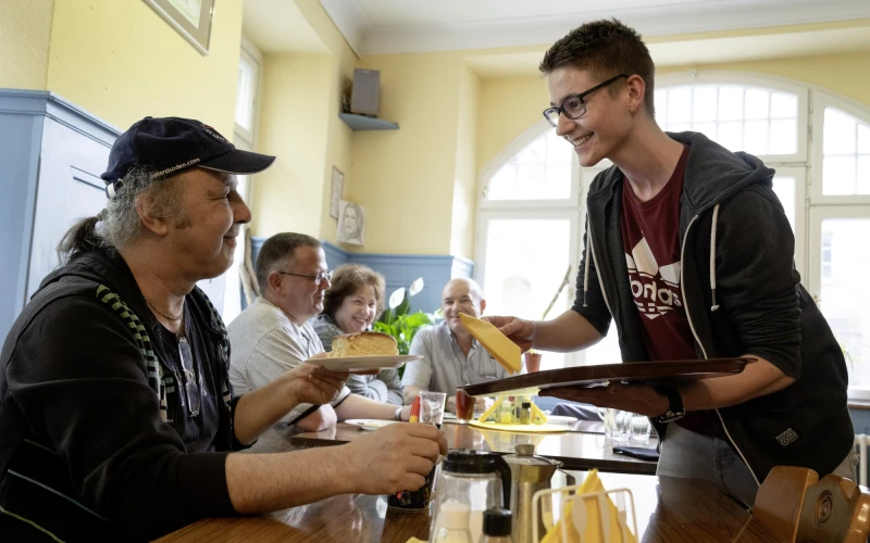 Junger Mann serviert Kuchen und Getr&auml;nk an einen &auml;lteren Mann an einem Tisch in einem hellen Raum, w&auml;hrend im Hintergrund weitere Personen sitzen und Mahlzeiten einnehmen.