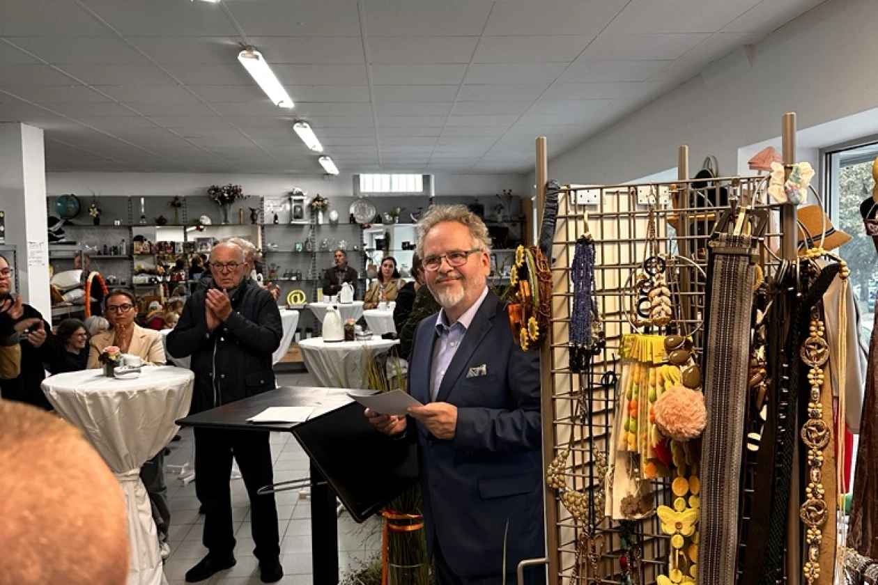 Ein grauhaariger Mann mit Bart und Brille steht in einem Laden an einem Rednerpult inmitten von Pflanzendeko, Schmuckauslage und Stehtischen. Er l&auml;chelt in die Kamera und scheint mit Menschen zu sprechen.