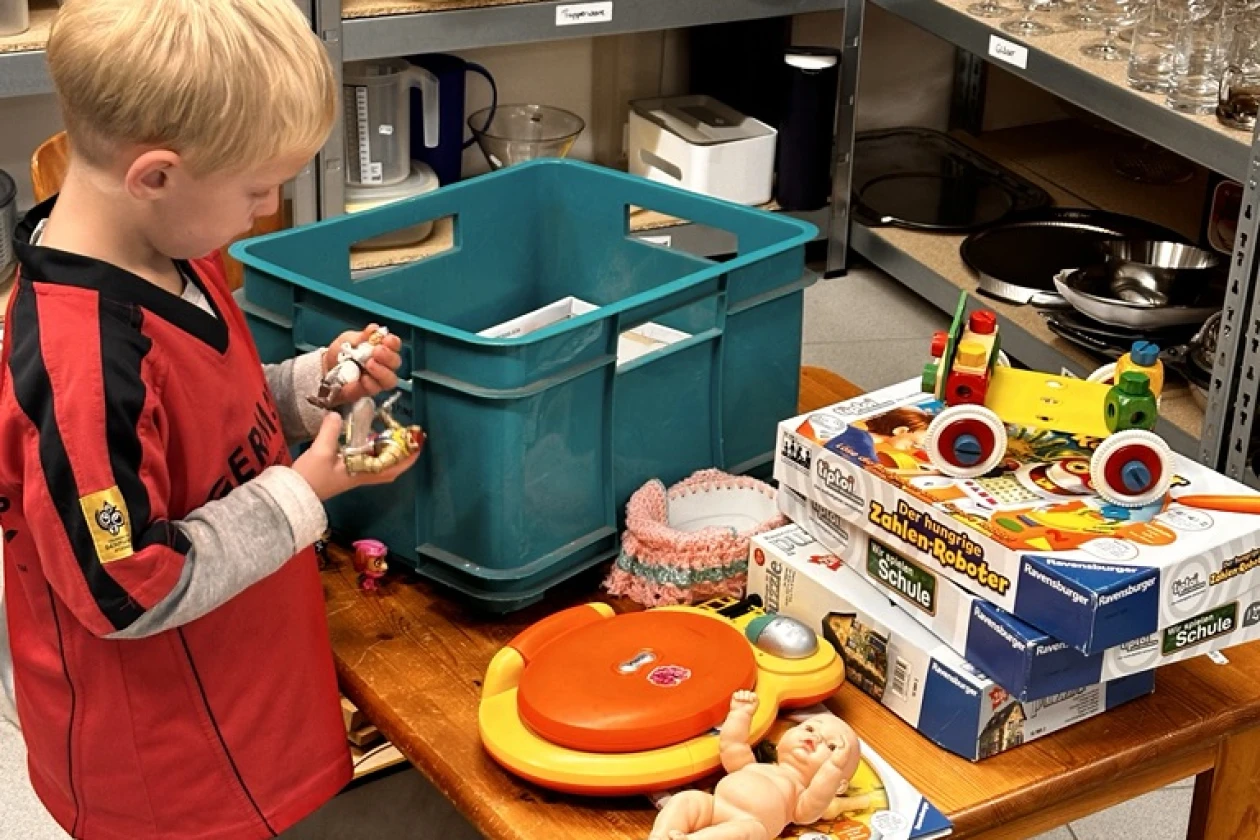 Ein kleiner Junge mit blonden Haaren in einem roten Shirt steht an einem Holztisch zwischen Regalen in einem Laden und spielt neben verschiedenen Kisten, Geschirr und Spielzeugkartons.