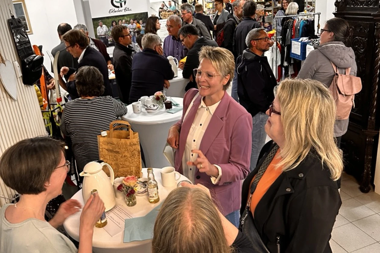 Gesch&auml;ftige Veranstaltung in einem Laden mit vielen G&auml;sten, die miteinander an Stehtischen und im Raum verteilt interagieren, w&auml;hrend eine Frau in einem rosa Blazer im Vordergrund steht.