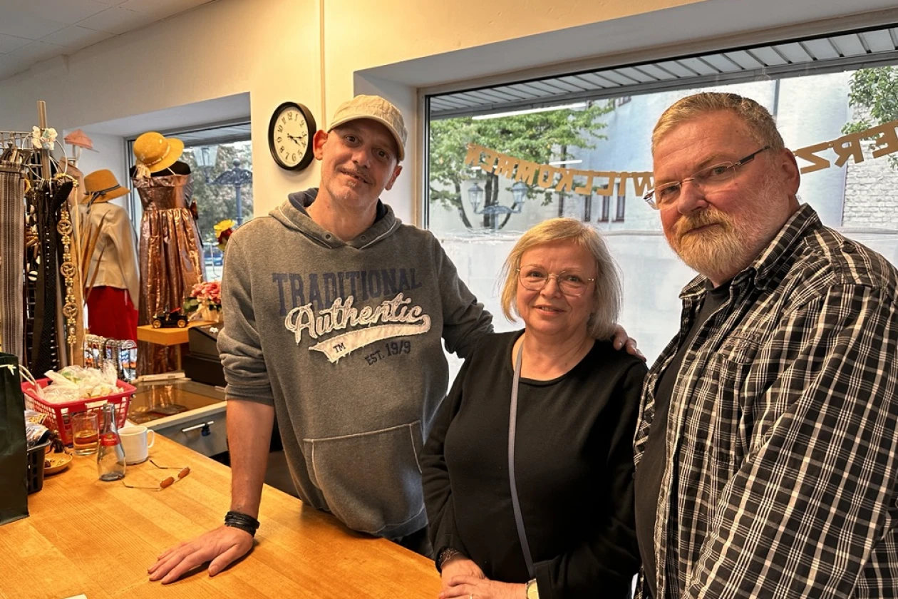 Eine Frau steht zwischen zwei M&auml;nnern hinter einer Theke, sie scheinen in einem Laden zu arbeiten. Alle drei l&auml;cheln in die Kamera. Im Hintergrund sind Schaufensterpuppen und Schmuckauslagen zu sehen.