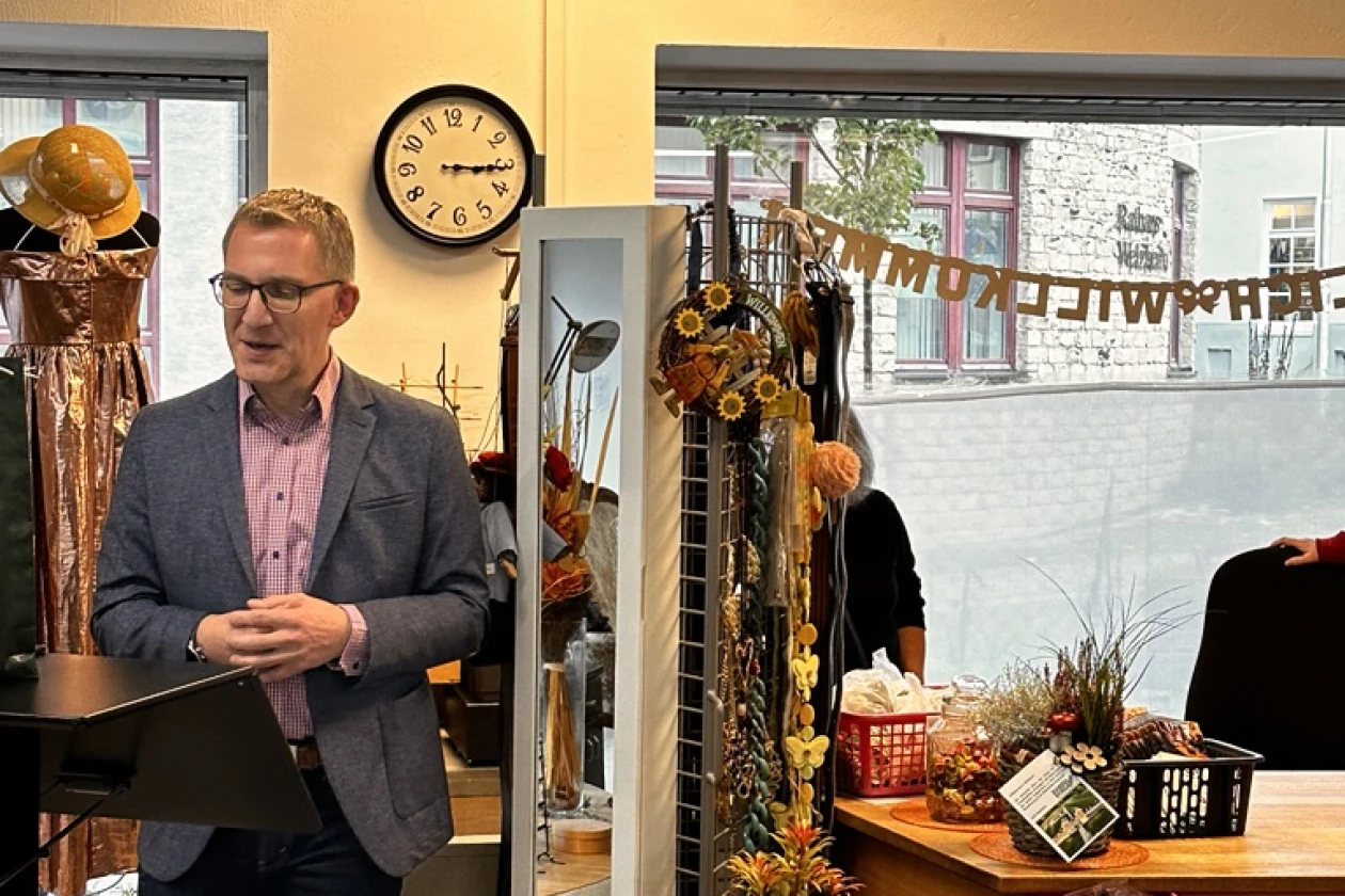 Ein Mann mit Brille steht an einem Rednerpult. Er hat seine H&auml;nde vor dem K&ouml;rper ineinandergelegt. Im Hintergrund ist ein dekoriertes Schaufenster und eine Ladentheke zu erkennen.