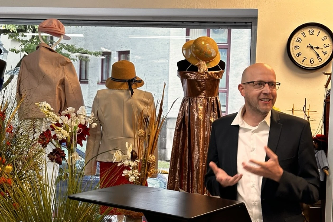 Ein Mann mit Brille steht an einem Rednerpult. Er gestikuliert mit seinen H&auml;nden. Im Hintergrund sind Pflanzendeko und Schaufensterpuppen in einem Schaufenster zu sehen.
