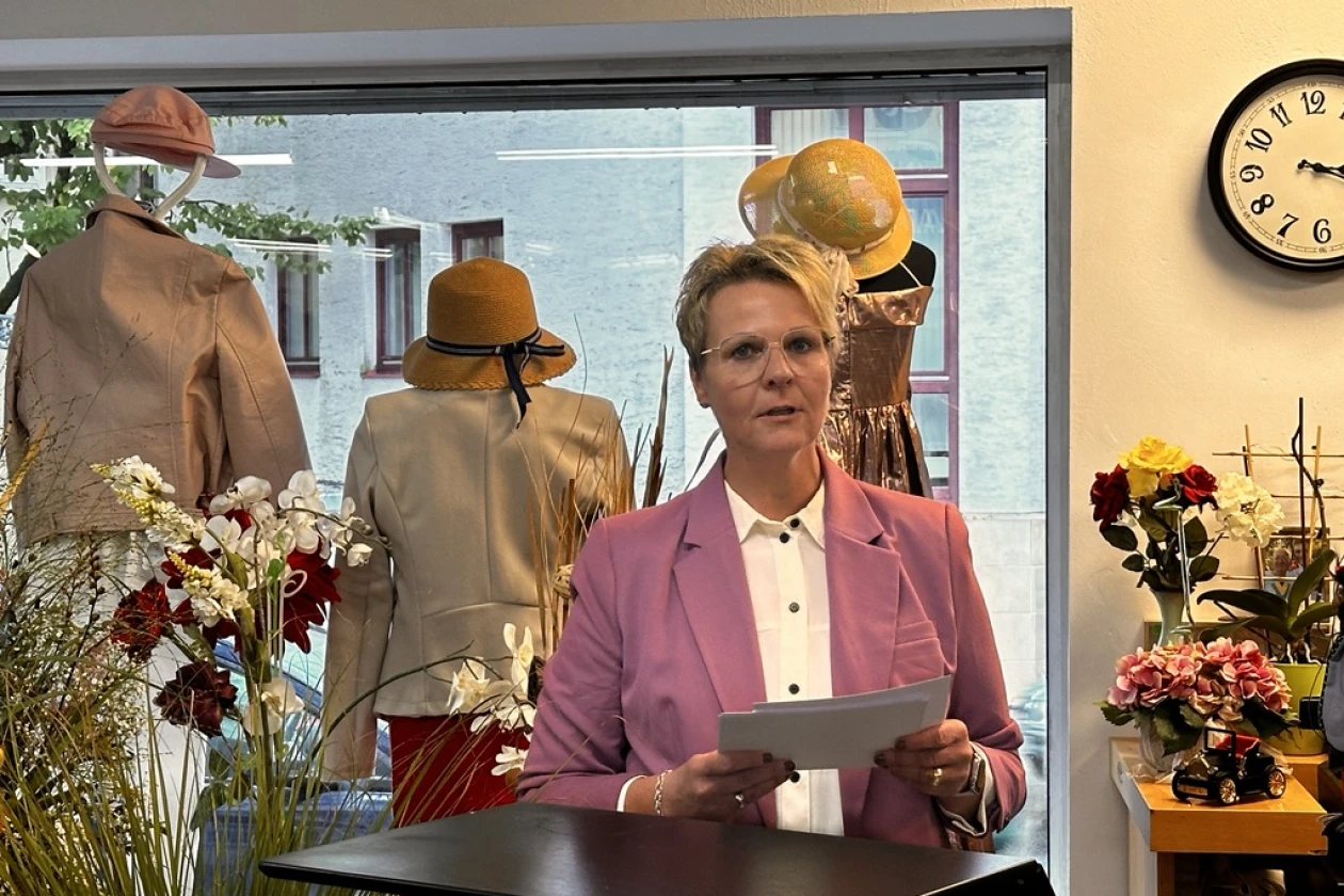 Eine blondhaarige Frau mit rosa Blazer steht an einem Rednerpult. Sie hat ihr Manuskript in den H&auml;nden. Im Hintergrund sind Pflanzendeko und Schaufensterpuppen in einem Schaufenster zu sehen.