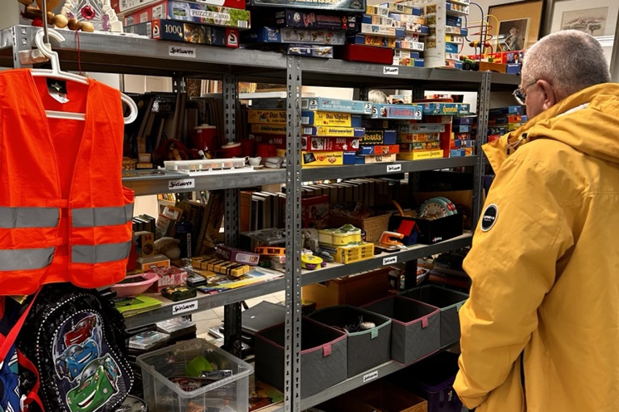 &Auml;lterer Mann in gelber Jacke steht neben Regalen voller Brettspiele, Spielzeug und Zubeh&ouml;r in einem Lagerraum mit roter Sicherheitsweste im Vordergrund.