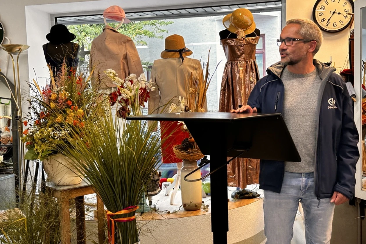 Ein Mann steht an einem Rednerpult in einem Laden. Er tr&auml;gt eine blaue Jacke mit einem Logo der Erlacher H&ouml;he. Im Hintergrund sind Pflanzendekoration und Schaufensterpuppen zu sehen.