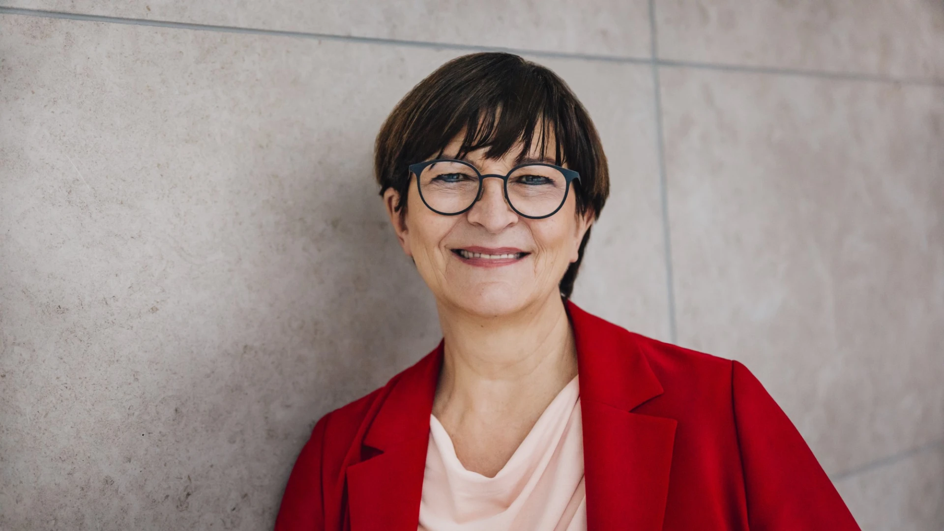 L&auml;chelnde Person mit kurzen dunklen Haaren und Brille, tr&auml;gt eine rote Jacke &uuml;ber einem rosafarbenen Oberteil. Sie steht vor einer neutralen grauen Wand und strahlt freundlich in die Kamera.
