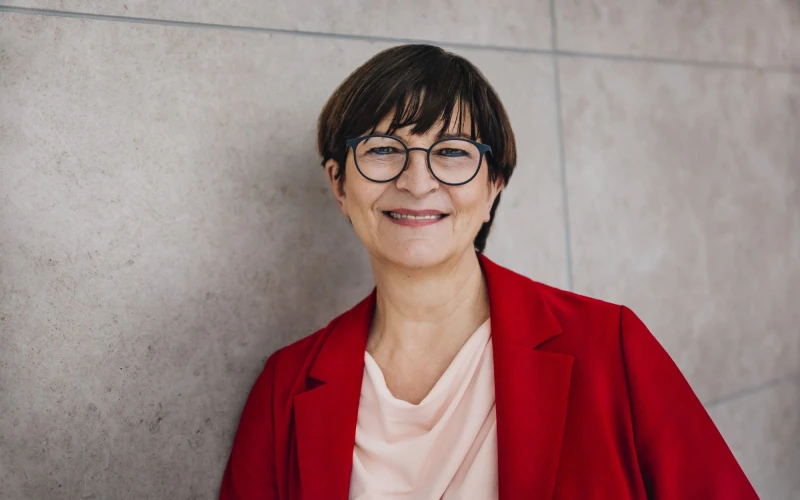 L&auml;chelnde Person mit kurzen dunklen Haaren und Brille, tr&auml;gt eine rote Jacke &uuml;ber einem rosafarbenen Oberteil. Sie steht vor einer neutralen grauen Wand und strahlt freundlich in die Kamera.