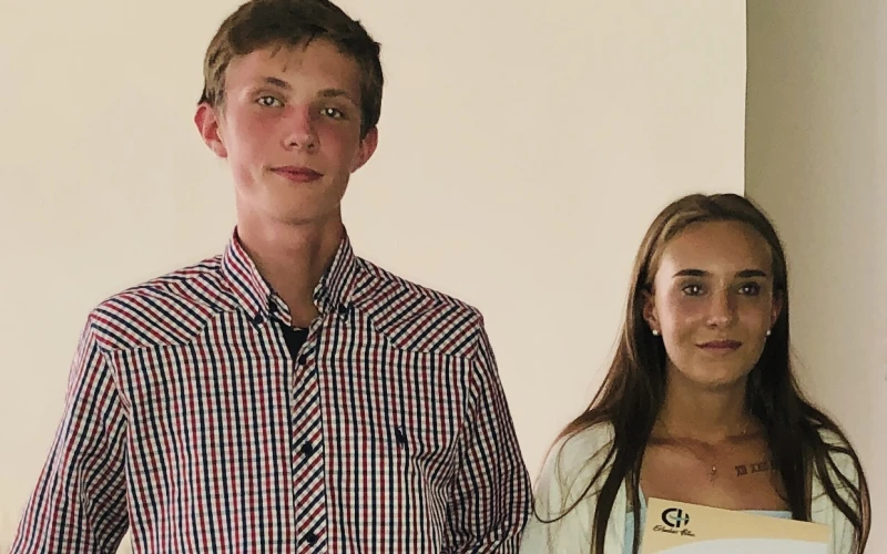 Zu sehen ist links ein Junge in einem karierten Hemd, rechts ein M&auml;dchen in einer wei&szlig;en Jacke. Beide halten ein Dokument in der Hand, das mit dem Logo der Erlacher H&ouml;he versehen ist.