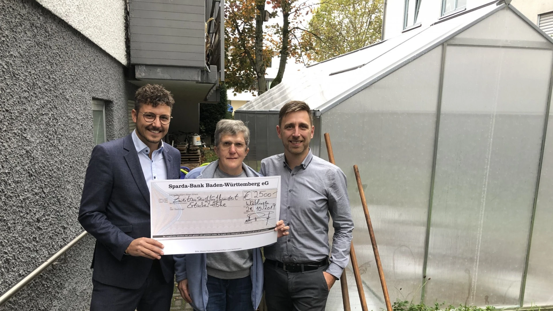 Drei Personen stehen vor einem Gew&auml;chshaus. Die Frau in der Mitte h&auml;lt gemeinsam mit dem Mann links einen SpendenScheck in der Hand . Im Hintergrund ist eine graue Hausfassade zu sehen.