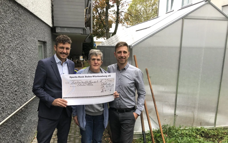 Drei Personen stehen vor einem Gew&auml;chshaus. Die Frau in der Mitte h&auml;lt gemeinsam mit dem Mann links einen SpendenScheck in der Hand . Im Hintergrund ist eine graue Hausfassade zu sehen.