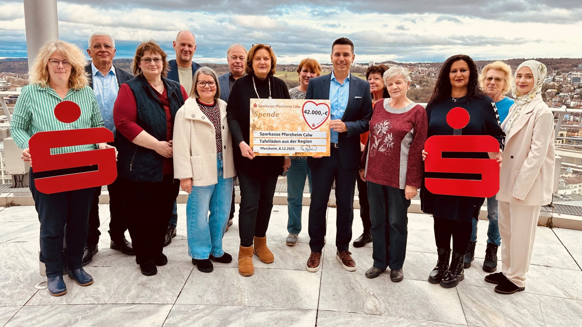 Eine Gruppe von etwa 15 Personen steht auf einer Aussichtsplattform hoch &uuml;ber einer Stadt und h&auml;lt einen gro&szlig;en Spendenscheck in die Kamera