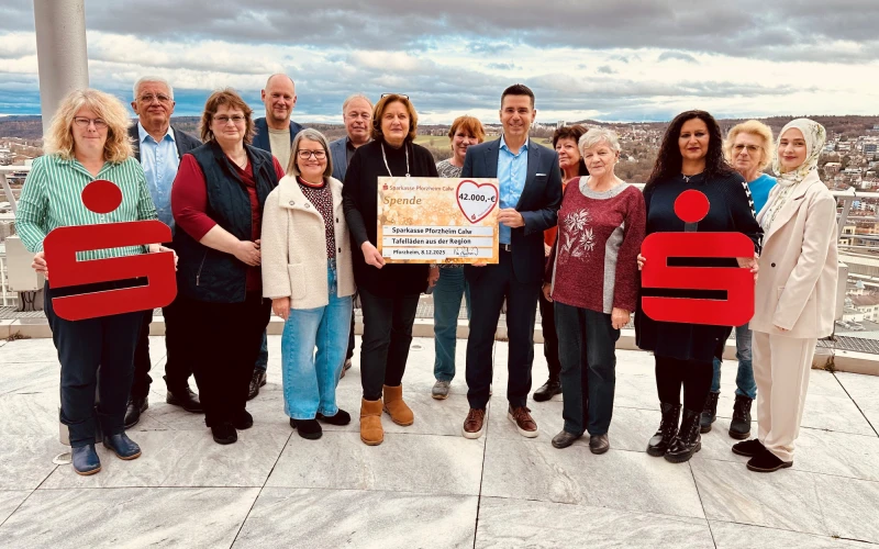 Eine Gruppe von etwa 15 Personen steht auf einer Aussichtsplattform hoch &uuml;ber einer Stadt und h&auml;lt einen gro&szlig;en Spendenscheck in die Kamera