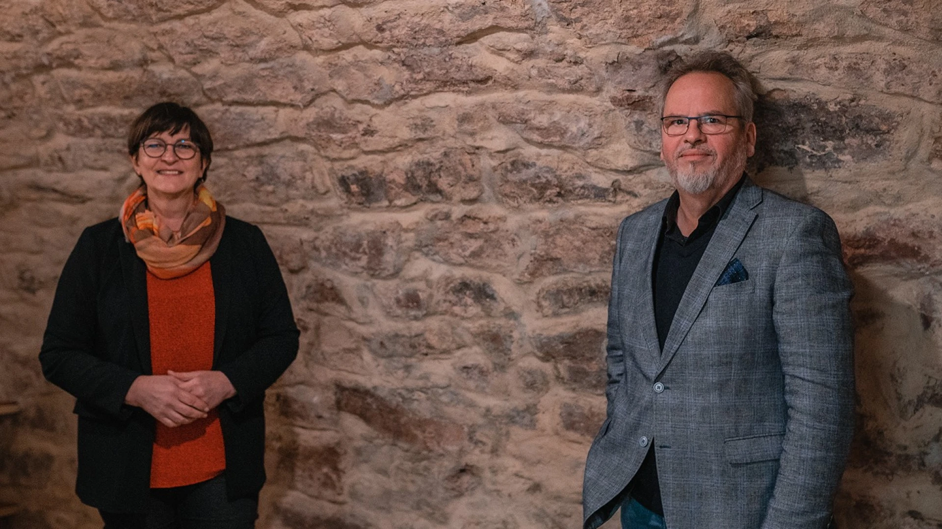 Eine Frau und ein Mann stehen in einem Gew&ouml;lbekeller vor einer Wand. Die Frau auf der linken Seite tr&auml;gt einen Schal, eine Brille, ein rotes Oberteil und eine schwarze Jacke dar&uuml;ber. Der Mann rechts tr&auml;gt ebenfalls eine Brille, ein schwarzes Hemd und dar&uuml;ber ein graues Jackett.