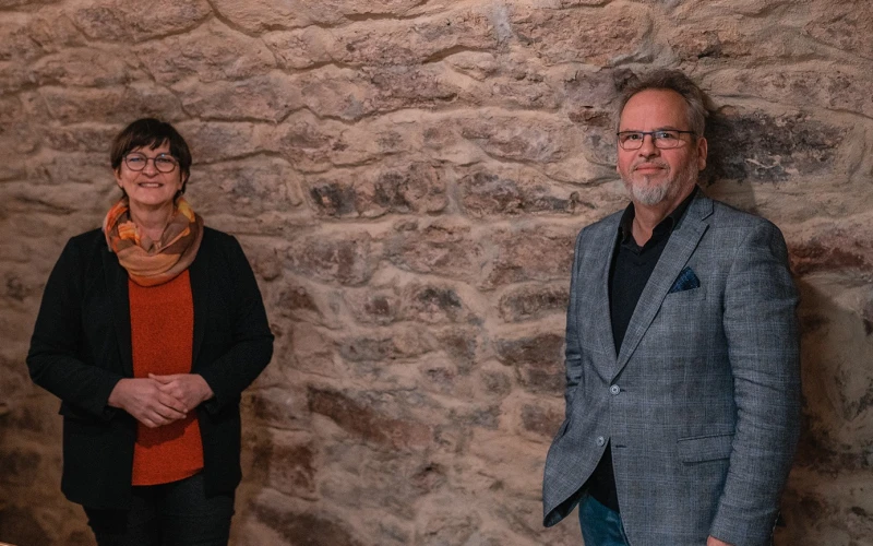 Eine Frau und ein Mann stehen in einem Gew&ouml;lbekeller vor einer Wand. Die Frau auf der linken Seite tr&auml;gt einen Schal, eine Brille, ein rotes Oberteil und eine schwarze Jacke dar&uuml;ber. Der Mann rechts tr&auml;gt ebenfalls eine Brille, ein schwarzes Hemd und dar&uuml;ber ein graues Jackett.