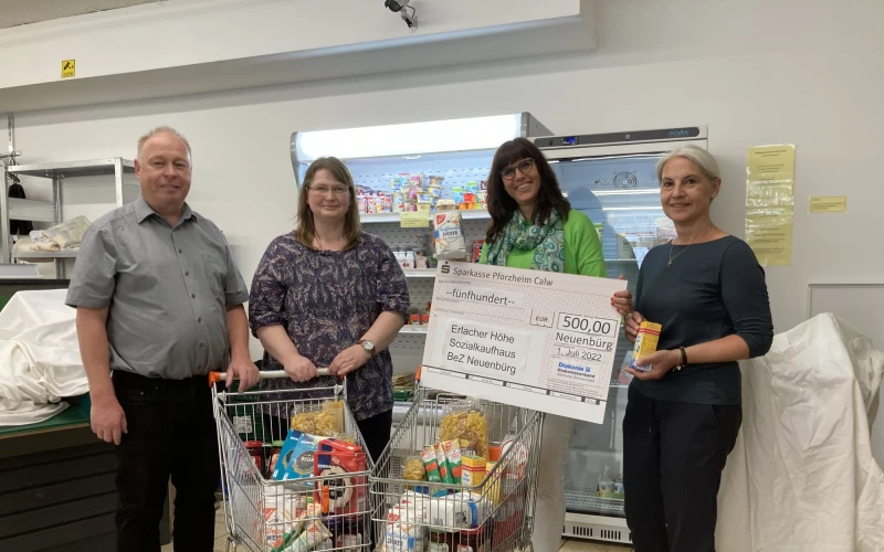 Drei Frauen und ein Mann vor K&uuml;hlregalen und gef&uuml;llten Einkaufsw&auml;gen vor einem K&uuml;hlregal. Ein Spendenscheck wird &uuml;bergeben. Alle l&auml;cheln freundlich in die Kamera.