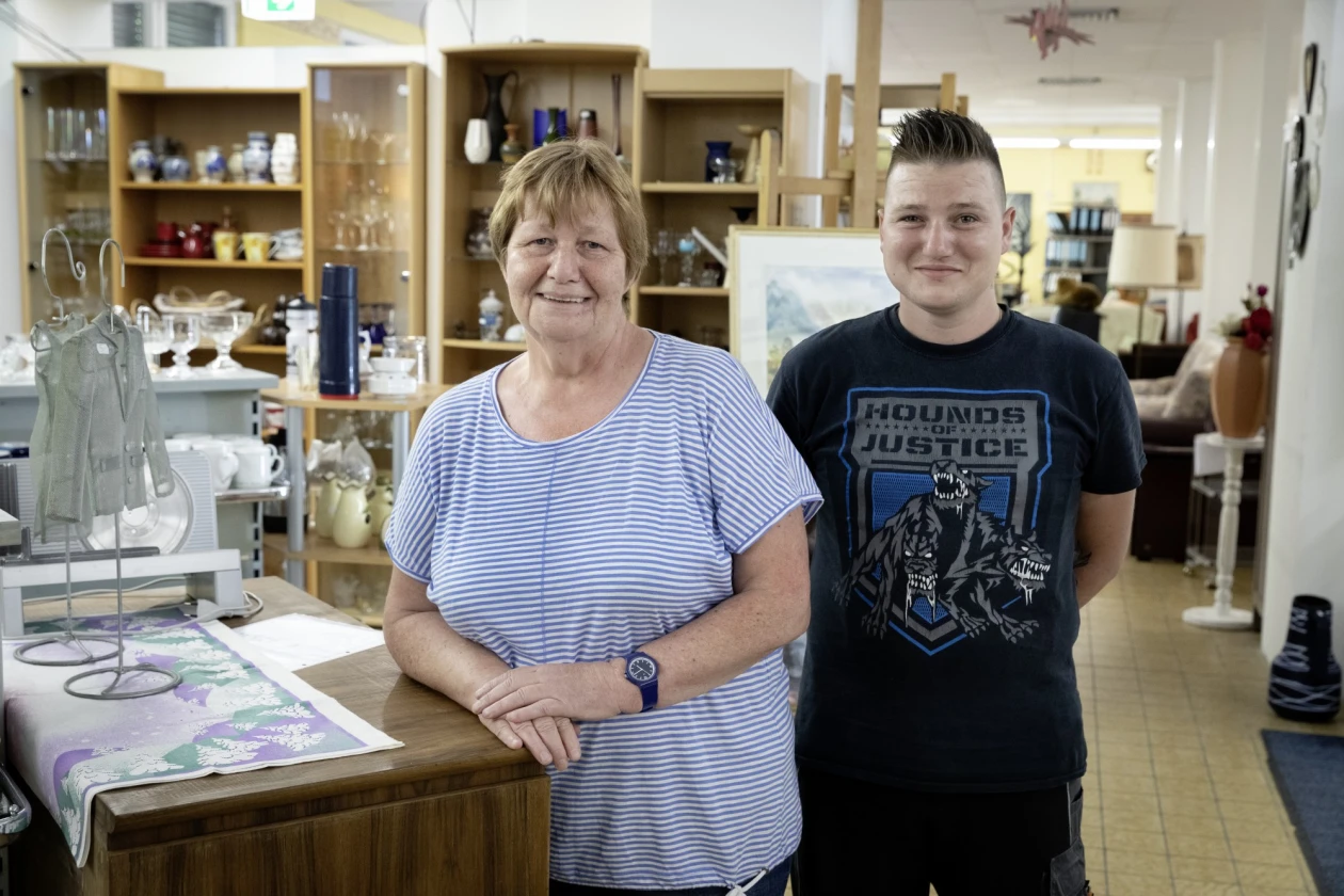 Zwei Personen stehen in einem Raum mit Regalen voller Geschirr, Gl&auml;ser und anderen Haushaltsgegenst&auml;nde. Die Frau links lehnt sich leicht an eine Theke. Beide l&auml;cheln in die Kamera.