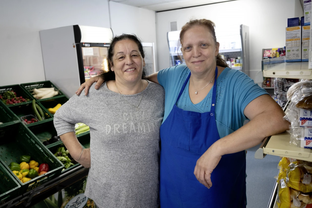 Zwei Frauen stehen in einem Gesch&auml;ft nebeneinander. Die Frau links tr&auml;gt ein graues Oberteil und die Frau rechts ein hellblaues T-Shirt mit blauer Sch&uuml;rze. Im Hintergrund sind Regale mit Lebensmittel, sowie Kisten mit Obst und Gem&uuml;se zu sehen.