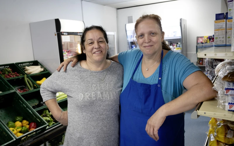 Zwei Frauen stehen in einem Gesch&auml;ft nebeneinander. Die Frau links tr&auml;gt ein graues Oberteil und die Frau rechts ein hellblaues T-Shirt mit blauer Sch&uuml;rze. Im Hintergrund sind Regale mit Lebensmittel, sowie Kisten mit Obst und Gem&uuml;se zu sehen.