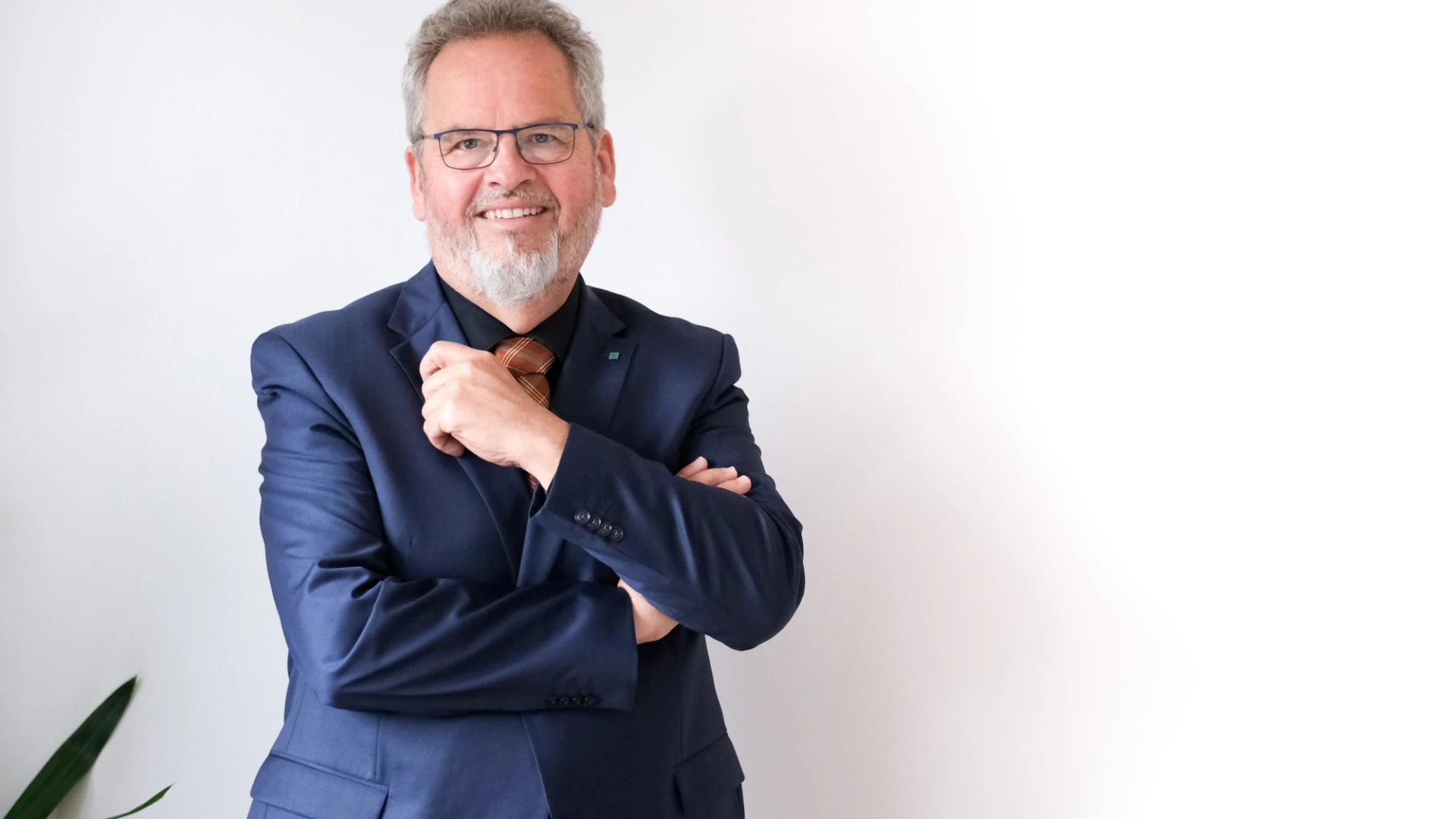 Ein &auml;lterer Mann im dunkelblauen Anzug mit Brille und grauem Bart l&auml;chelt in die Kamera. Er pr&auml;sentiert sich selbstbewusst mit verschr&auml;nkten Armen vor dem K&ouml;rper.