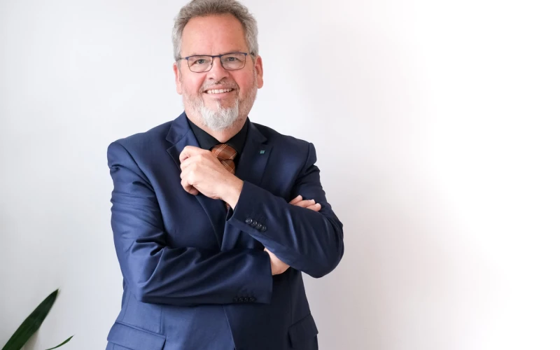 Ein &auml;lterer Mann im dunkelblauen Anzug mit Brille und grauem Bart l&auml;chelt in die Kamera. Er pr&auml;sentiert sich selbstbewusst mit verschr&auml;nkten Armen vor dem K&ouml;rper.