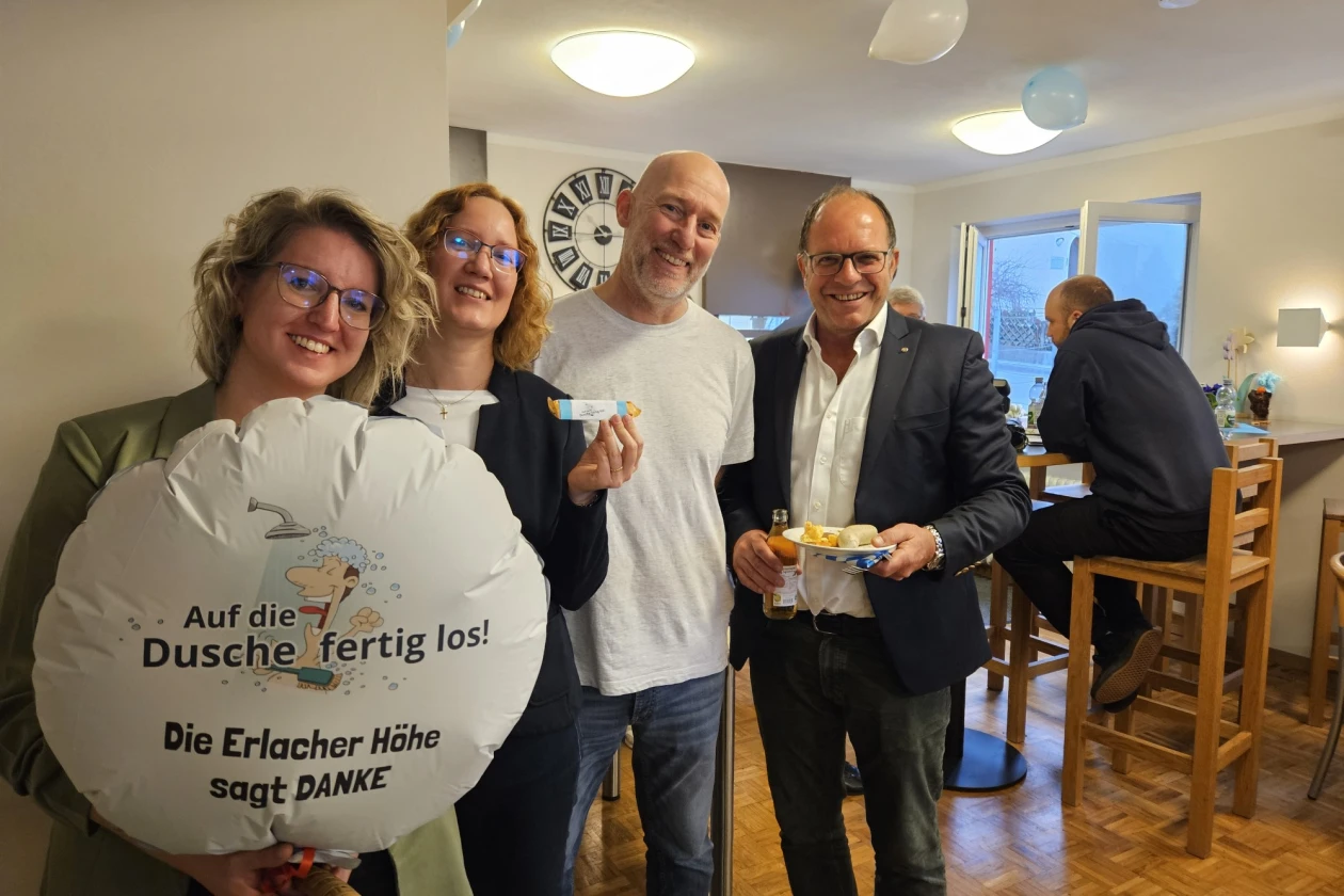 Vier Personen stehen hintereinander in einem Raum, zwei Frauen und zwei M&auml;nner, in lockerer Atmosph&auml;re. Eine Frau h&auml;lt einen runden Lufballon mit der Aufschrift "Auf die Dusche fertig los! Die Erlacher H&ouml;he sagt DANKE". Die Gruppe scheint in einem Bistroraum zu sein, mit Holzm&ouml;beln und Beleuchtung im Hintergrund.